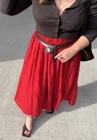 Osoba koja nosi crnu bluzu na dugmad i crvenu suknju s smešnim pojasom, drži pojas jednom rukom i stoji na betonu.