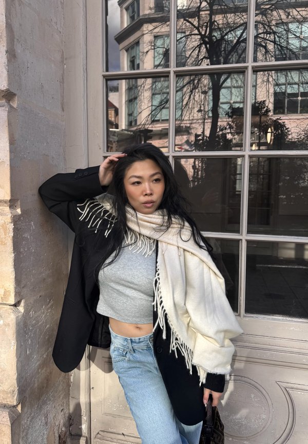 Femme aux cheveux longs et noirs appuyée contre un mur en pierre, portant un blazer noir, une écharpe blanche à franges, un crop top gris, un jean clair, tenant un petit sac.