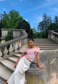 Giovane donna in top rosa senza spalline e gonna bianca appoggiata a un parapetto di pietra accanto a una scala in cemento all'aperto, con alberi e cielo blu sullo sfondo.