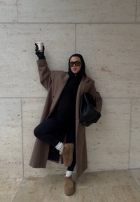 Personne en manteau marron, sweat à capuche noir, lunettes de soleil, gants, et bottes beige posant avec une jambe levée, tenant une tasse de café et un sac noir près d'un mur en pierre.