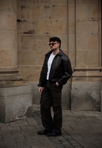 Schwarze Lederjacke mit Reißverschluss, weißes T-Shirt, braune Cargo-Hose mit Taschen, schwarze Schuhe. Stehend vor einer Steinmauer, lässiger Streetstyle.