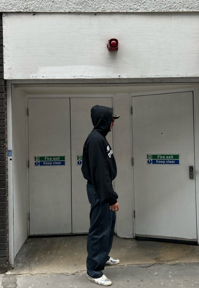Sudadera negra con texto blanco, jeans azules y zapatillas plateadas. El fondo presenta dos puertas blancas con señales de salida de emergencia verdes y una alarma roja.
