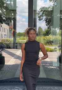 Μαύρο αμάνικο φόρεμα με υψηλό λαιμό και ραφές. Χρυσά κοσμήματα που προσδίδουν λάμψη, σε φόντο γυάλινου κτηρίου και καταπράσινης βλάστησης.