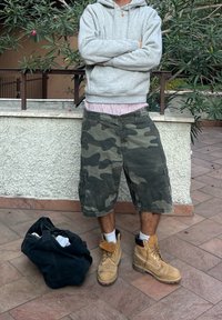 Hanorac gri cu glugă, pantaloni scurți cargo cu model de camuflaj, cizme de lucru maro. Geantă neagră pe jos. Setare în aer liber cu vegetație verde și podea din plăci.