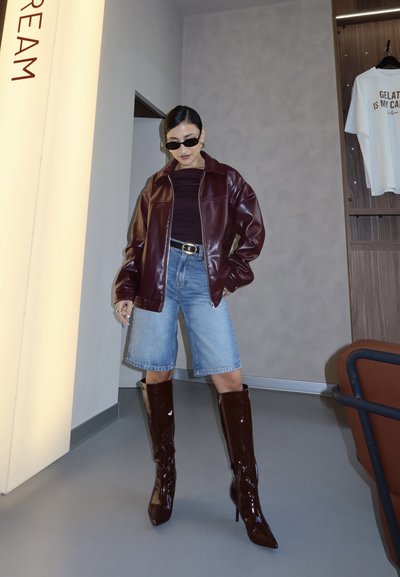 Mujer con gafas de sol, chaqueta de cuero burdeos, top negro, pantalones cortos vaqueros y botas brillantes hasta la rodilla en interior cerca de una pared y una camiseta colgada.