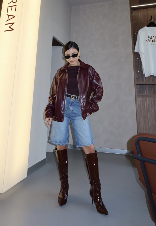 Žena v slnečných okuliaroch, burgundskej koženej bunde, čiernom topán, rifľových krátkych nohaviciach a lesklých čižmách po kolená vo vnútri blízko steny a zaveseného trička.