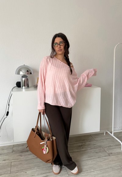 Mujer con gafas que lleva un suéter rosa holgado y pantalones negros, sosteniendo un bolso grande marrón, de pie en una habitación minimalista.