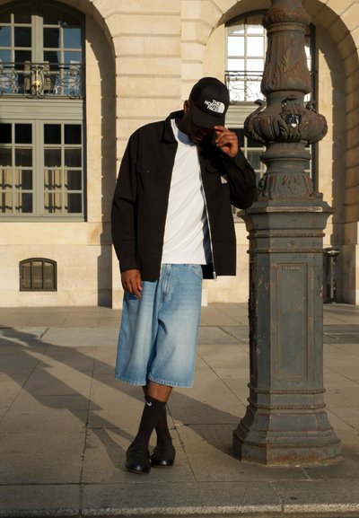 Chaqueta ligera negra sobre una camisa blanca, combinado con pantalones cortos de mezclilla azules. Gorra negra y calcetines oscuros con zapatos negros de tipo slip-on. Poste de lámpara decorativo cercano.