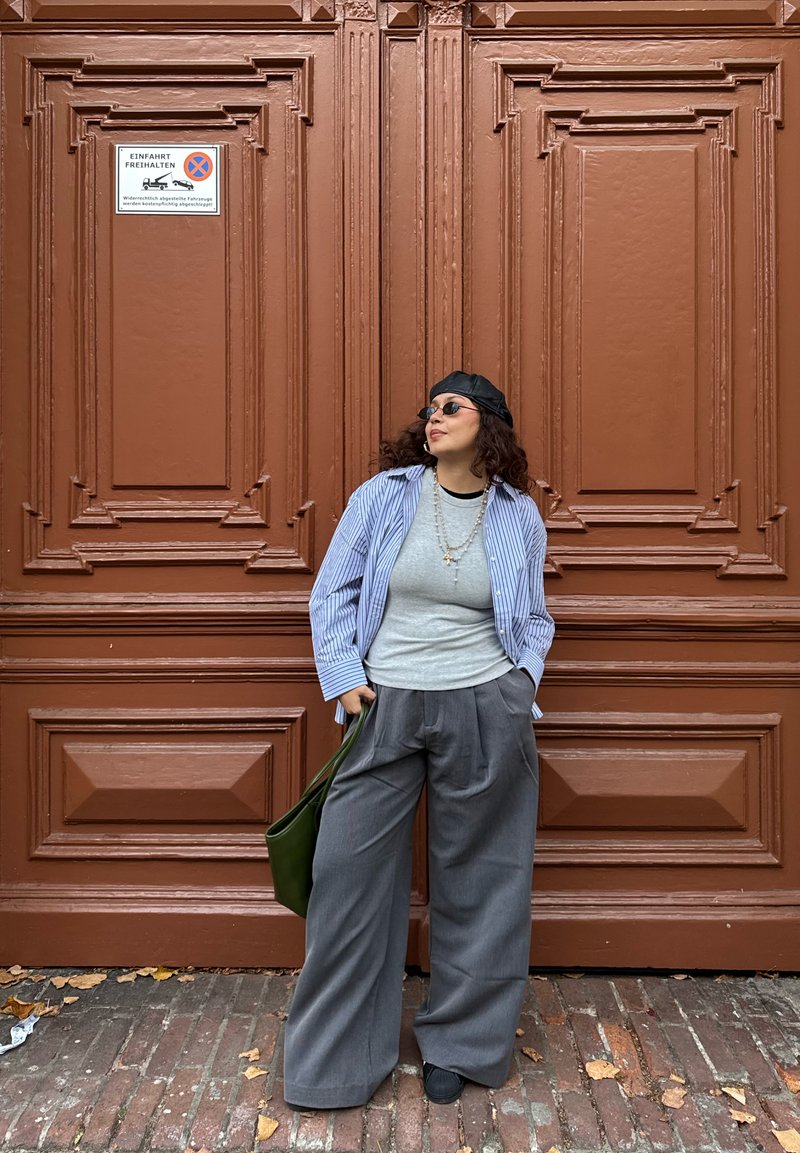 Chemise à rayures boutonnée, t-shirt gris, pantalon large gris, chaussures noires, sac vert. Fond en bois brun avec des détails ornés.