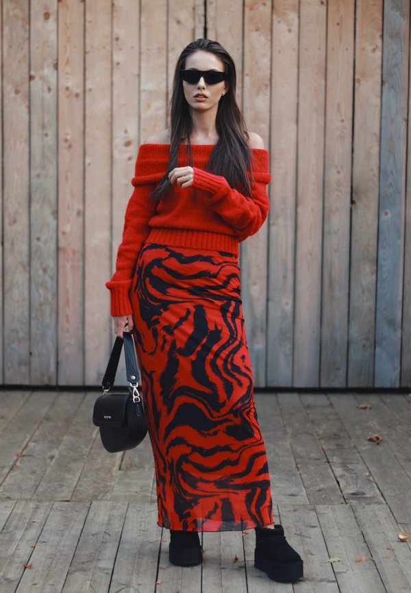 Rdeča majica z off-shoulder izrezom in maksi krilo z črno-rdečimi tigrovimi črtami. Model drži črno torbico in nosi črne sončne očala. Lesena ozadja.