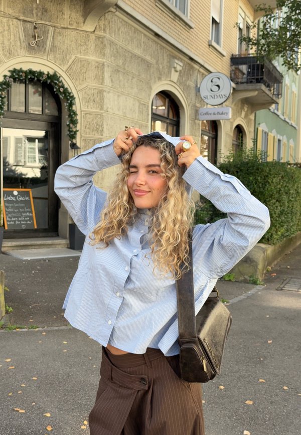 Ung kvinde med krøllet blondt hår iført en striped skjorte og brune bukser smiler skævt, mens hun justerer sin hætte uden for en café.