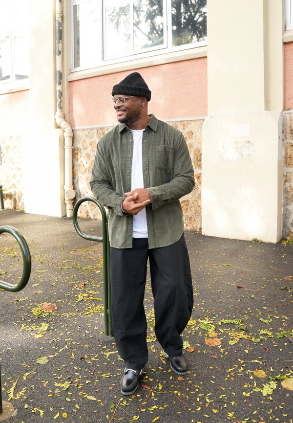 Un homme souriant, coiffé d’un bonnet noir, portant des lunettes et des vêtements décontractés, se tient dehors à côté d’un bâtiment, les mains jointes. Des feuilles jaunes recouvrent le trottoir.