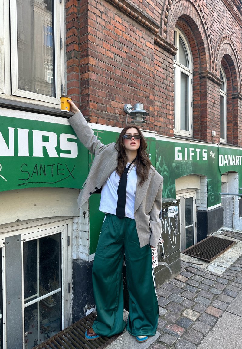 Young woman wearing oversized blazer, green wide pants, and sunglasses leans against brick building, holding a yellow coffee cup up near a window.
