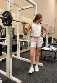 Barra con pesas en un soporte para sentadillas, persona vestida con una camiseta corta gris y pantalones cortos con un patrón y un cordón naranja, zapatillas blancas. Escenario de gimnasio.