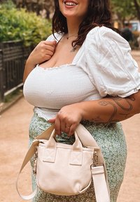 Pequena bolsa bege com duas alças e uma alça destacável, apresentando uma textura suave e fecho em zíper. Cercada por vegetação.