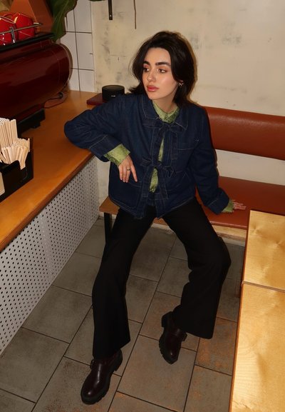 Chaqueta de denim azul oscuro con cierre de lazo, camisa verde con cuello debajo, pantalones negros acampanados y botas burdeos. Ambiente simple de café.