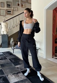 Γυναίκα με μαύρο φόρμα, γκρι αθλητικό σουτιέν και μαύρο μπουφάν που φοριέται μερικώς, στέκεται πάνω σε μαύρο και γκρι χαλί σε ένα μοντέρνο σαλόνι.