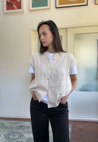 Vesta bej din in, cu mâneci scurte, închidere cu nasturi, croială lejeră, purtată peste un tricou alb, asortată cu pantaloni negri. Design simplu, textură ușoară.
