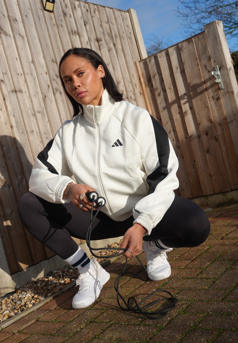 Femme en veste de sport blanche et noire accroupie sur une terrasse en briques, tenant une corde à sauter noire, avec une clôture en bois et un ciel dégagé en arrière-plan.