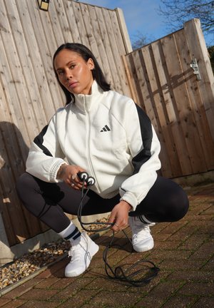 Mujer con chaqueta deportiva blanca y negra en cuclillas sobre un patio de ladrillos, sosteniendo una cuerda para saltar negra, con una cerca de madera y cielo despejado de fondo.