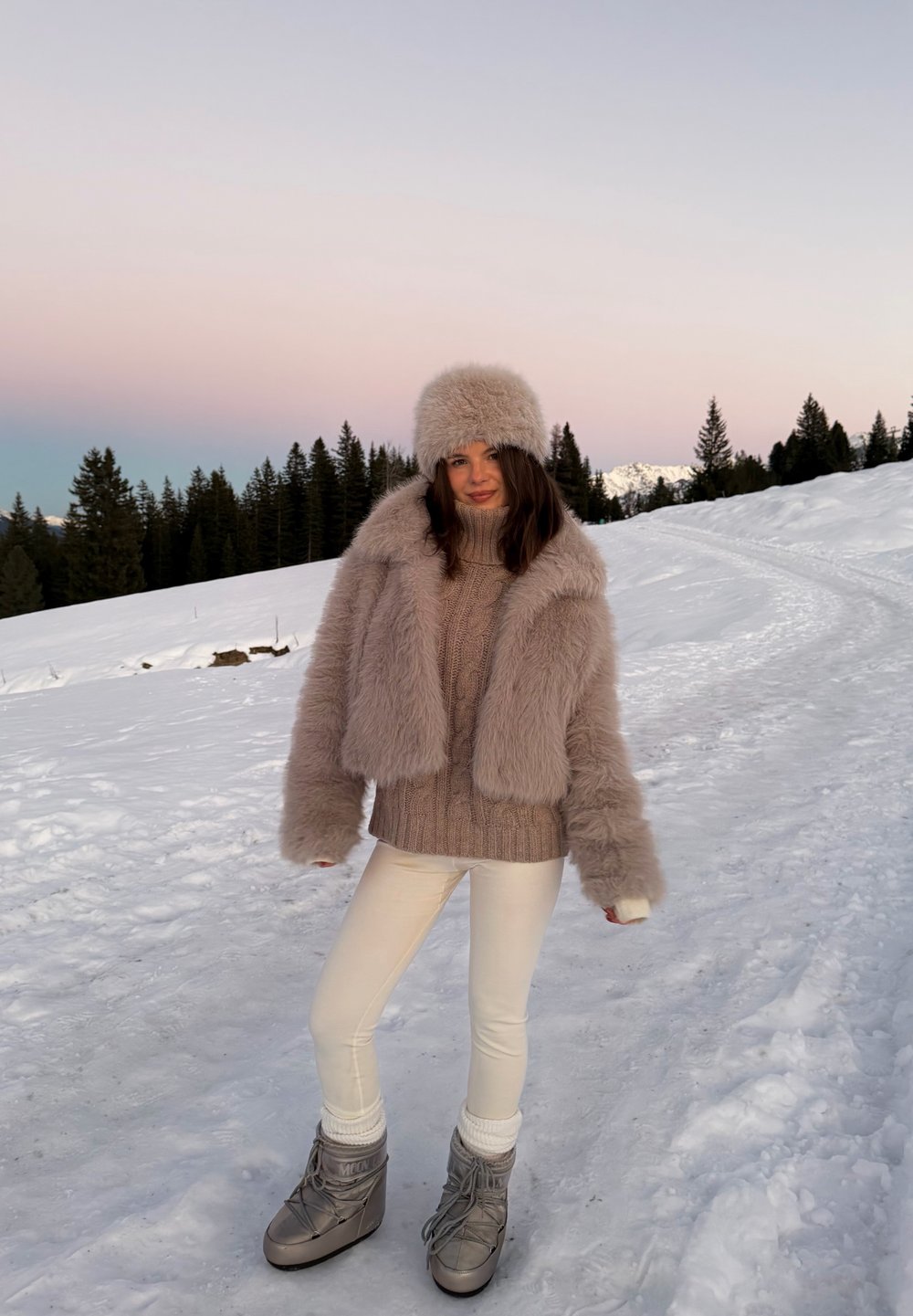 Veste en fausse fourrure beige clair à manches larges, assortie à un pull beige en maille, des leggings blancs et des bottes d'hiver grises, sur fond de neige.