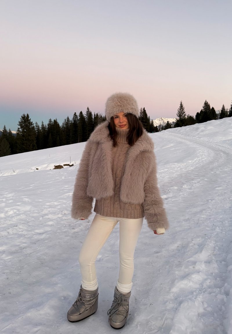 Veste en fausse fourrure beige clair à manches larges, assortie à un pull beige en maille, des leggings blancs et des bottes d'hiver grises, sur fond de neige.