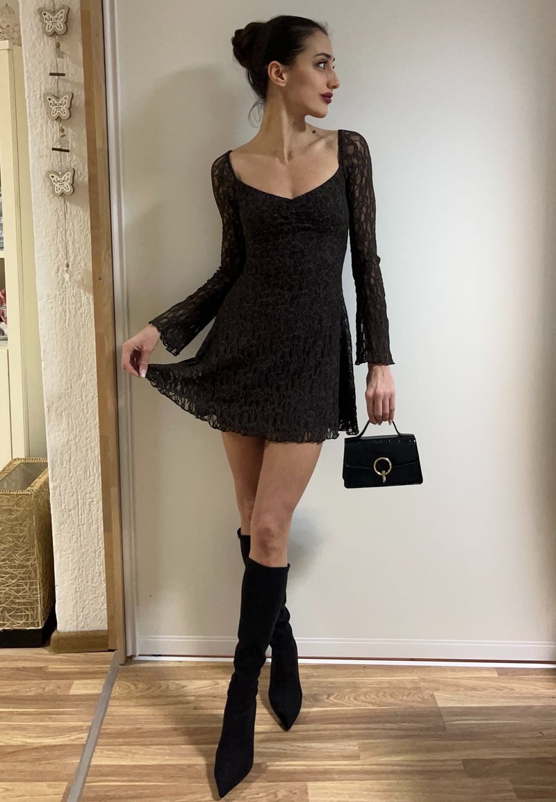 Γυναίκα με μαύρο μίνι φόρεμα από δαντέλα με μακριά μανίκια, κρατώντας μικρή μαύρη τσάντα χειρός, φοράει μαύρες μπότες μέχρι το γόνατο, στέκεται σε ξύλινο πάτωμα.