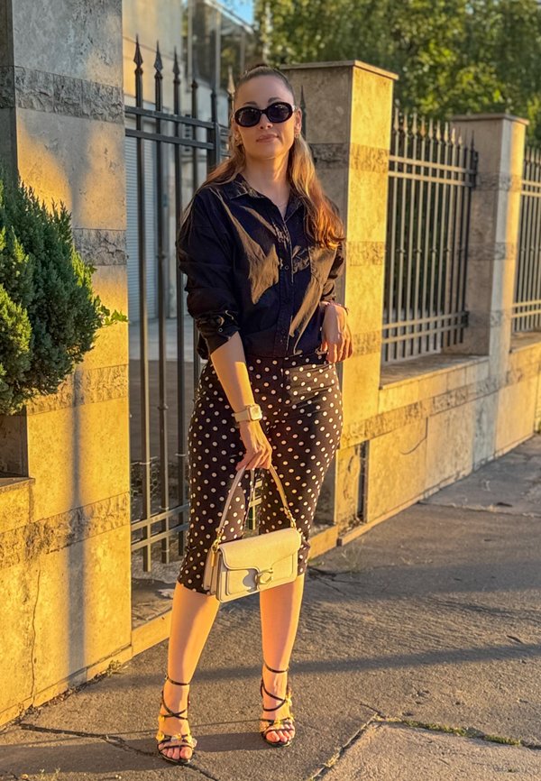 Vrouw met zwart shirt, gestippelde rok, zonnebril, beige handtas in haar hand, staand bij een stenen muur en een metalen hek in het zonlicht.