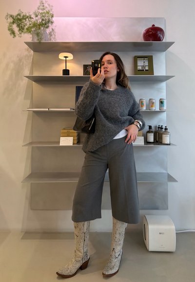 Mujer con suéter gris, pantalones de pierna ancha y botas blancas hasta la rodilla con estampado, tomando una selfie frente al espejo en una habitación moderna con estanterías.