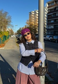 Vestă din denim peste o cămașă cu dungi, pantaloni roz cu croială largă, șapcă mov, ochelari de soare și geantă maro purtată peste umăr. Cadru urban în aer liber cu copaci.