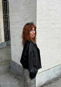 Zwarte leren jas over een patroon van leopardenprint rok, met lang rood haar en een zonnebril op het hoofd. Staand naast een gestructureerde betonnen muur.