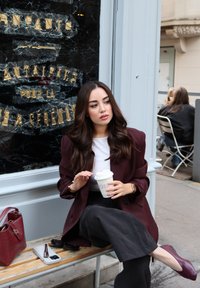 Blazer oversize color bordeaux sopra una maglietta bianca, pantaloni neri, mentre tiene in mano una tazza da caffè. Una borsa bordeaux e un paio di occhiali da sole riposano su una panchina di legno.