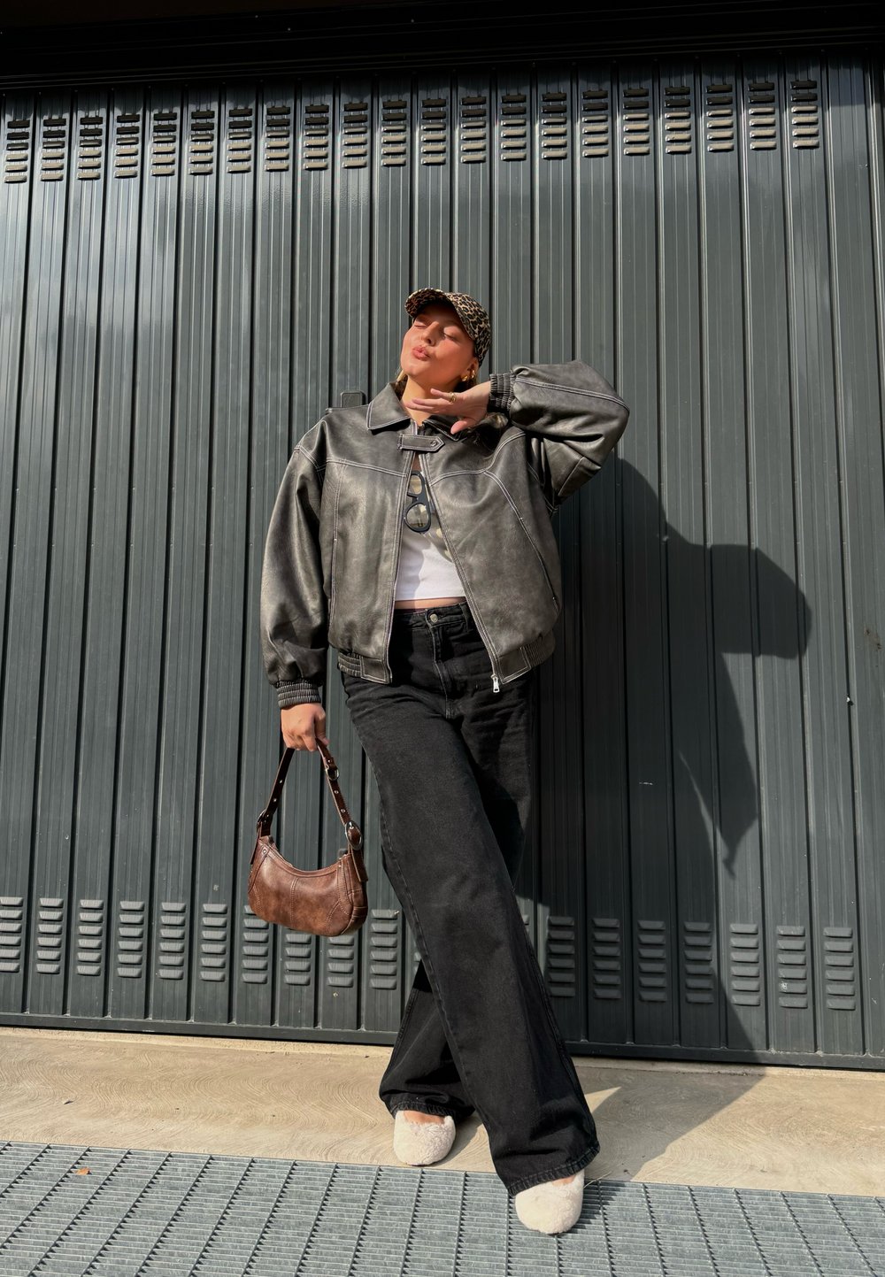 Graue Lederbomberjacke, weißes Oberteil, schwarze weitgeschnittene Hose, braune Handtasche, beiges, plüschiges Schuhwerk, steht vor einer dunklen wellenförmigen Wand.