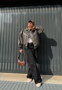 Graue Lederbomberjacke, weißes Oberteil, schwarze weitgeschnittene Hose, braune Handtasche, beiges, plüschiges Schuhwerk, steht vor einer dunklen wellenförmigen Wand.