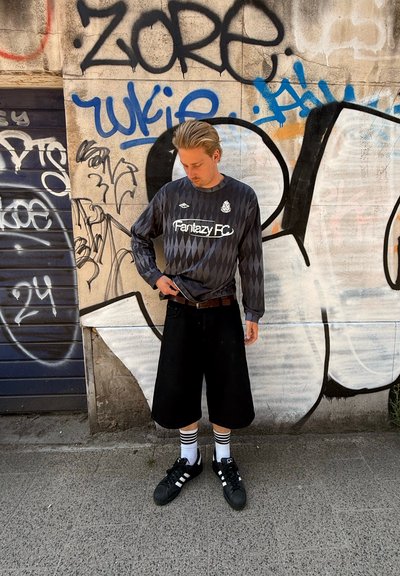 Jersey de manga larga en gris oscuro con patrón de diamantes y logo de "Fantazy FC", combinado con pantalones cortos negros hasta la rodilla y zapatillas negras de Adidas.