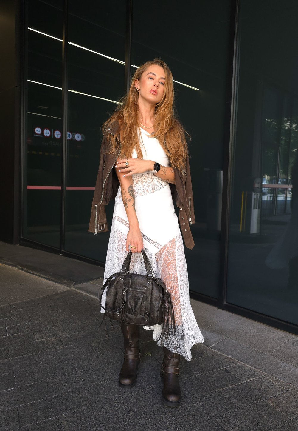 Brown leather jacket over a white top and a lace white skirt, matched with knee-high brown boots and a textured black bag.