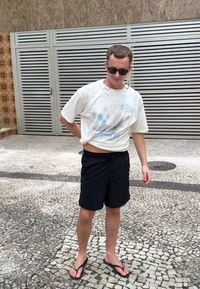 Joven con gafas de sol, camiseta blanca con diseño gráfico, pantalones cortos negros y chanclas, de pie sobre un pavimento de baldosas ajustándose la camisa.