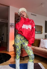 Personne portant une veste rouge Puma, un béret beige, des leggings verts à motifs et des bottes blanches, posant à l'intérieur près d'un canapé rose et d'un sol en bois.