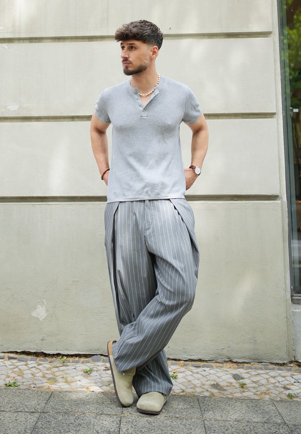 Homme aux cheveux courts et foncés, debout contre un mur, portant une chemise grise à manches courtes, un pantalon rayé à jambes larges, des chaussures beiges et un collier de perles.