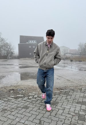 Chaqueta bomber gris, camisa rosa, vaqueros azules holgados y zapatillas brillantes rosas. Suelo pavimentado y un fondo brumoso con edificios.