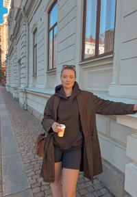 Um casaco castanho sobre um hoodie preto, combinado com calções pretos. Segurando uma chávena de café, parado ao lado de um edifício branco ornamentado numa rua de pedra.
