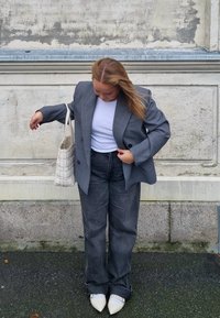 Sivi oversize sako preko bijele majice, uparen s tamnim trapericama i svijetlim đonom cipelama. Izgled upotpunjava svijetlo kremasta pletena torba.