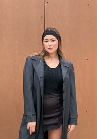 Μαύρο παλτό με μεγάλα κουμπιά πάνω από ένα πλεκτό μαύρο τοπ και μια κοντή μαύρη φούστα με δαντέλα στο τελείωμα. Το άτομο στέκεται μπροστά σε καφέ φόντο.