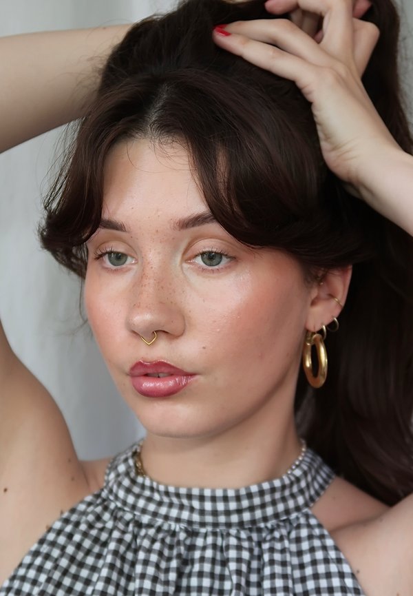 Giovane con pelle chiara, lentiggini, occhi verdi e capelli scuri, che indossa orecchini a cerchio dorati, anello al setto nasale e un top a quadretti bianco e nero.