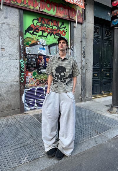 Camisa de manga corta a cuadros con un gráfico de calavera, combinada con pantalones de chándal gris de gran tamaño. Calzado negro, fondo de graffiti urbano.