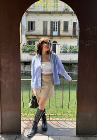 Ανοιχτό μπλε πουκάμισο με κουμπιά, λευκή τοπ crop, μπεζ μίνι φούστα, μαύρες μπότες μέχρι το γόνατο και ελιάς πράσινη τσάντα χειρός; στέκεται δίπλα σε μια πέτρινη περίφραξη.