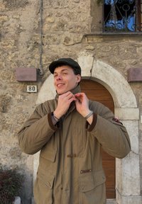 Jeune homme souriant en train de boutonner une veste marron, debout devant un mur en pierre avec une porte en bois arquée et une plaque numérotée 80.