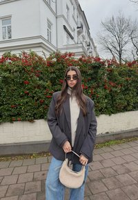 Femme aux longs cheveux bruns portant des lunettes de soleil, un blazer gris, un col roulé beige et un jean, tenant un sac à main beige, debout près de buissons verts avec des baies rouges.