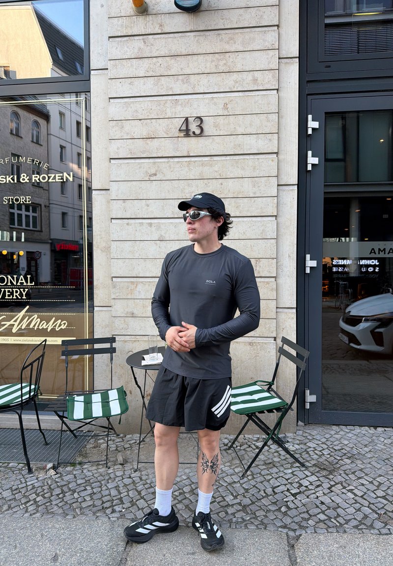 Homme portant une casquette noire, des lunettes de soleil, une chemise à manches longues et un short, debout devant un café avec des chaises rayées et un pavement en pavés.