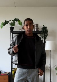 Veste en cuir noir avec un col doublé de fourrure, portée sur un sweat-shirt noir avec des accents rouges, associée à un pantalon gris clair à motifs.